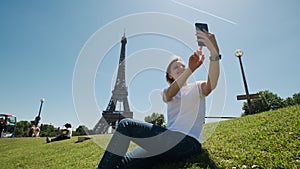Woman is taking selfie using smartphone sitting near Eiffel tower in Paris in daytime, Wide shot footage