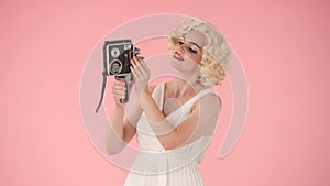 Woman taking selfies using old vintage video camera. Woman in look like Marilyn Monroe poses for camera on pink