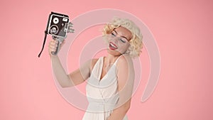 Woman taking selfies using old vintage video camera. Woman in look like Marilyn Monroe poses for camera on pink