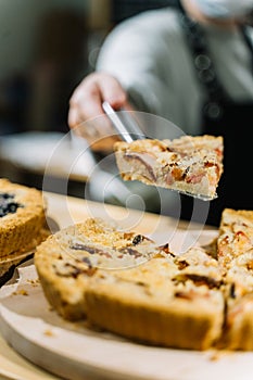 Woman taking spatula with piece of tasty open plum pie from wooden shelf with different kinds of pies at cafe.