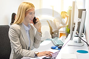 Woman talking on the mobile phone and smiling while sitting at her working place in office and looking at computer