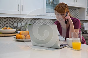 Woman talking on mobile phone while using laptop in kitchen