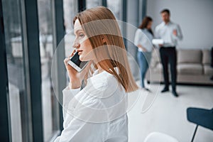 Woman talking by phone in front of group of young successful team that working and communicating together indoors in