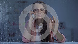 Woman talking phone and looking through rainy window, waiting husband at night