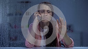 Woman talking phone and looking through rainy window, waiting husband at night