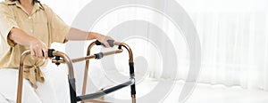A woman in a tan shirt is using a walker. The walker is made of metal and has a black handle, panorama concept