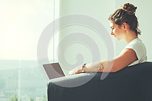 Woman with tattoo sitting with laptop in front of big bright window with panoramic view