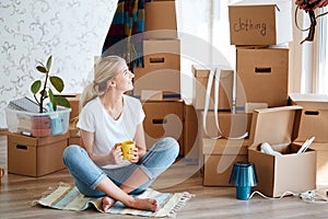 Woman with tea in hand sitting on floor of new apartment, pile of moving boxes on background