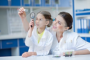 Woman teacher and girl student scientists looking at glass microscope slide through magnifier
