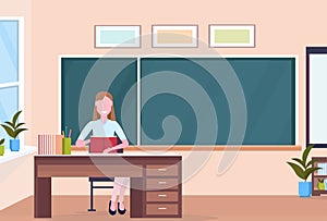 Woman teacher sitting at desk reading book education concept modern school classroom interior chalk board female cartoon