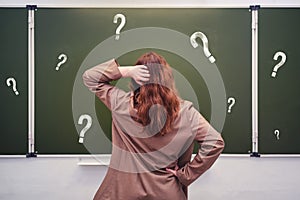Woman teacher stands pensively against the background of a school blackboard with question marks