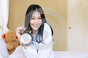 Woman with teddy bear holding alarm clock sitting on the white bed while looking at camera. Cheerful girl waking up