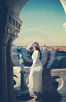 Woman with telescope at viewpoint on cityscape. Sensual woman with long hair in white wedding dress, fashion. Girl with