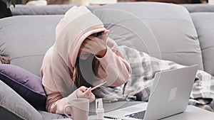 The woman with thermometer is lying in bed and looking for symptoms in laptop