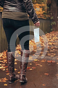 a woman in tight black jeans and boots holds a disposable protective face mask while walking in an urban surrounding.