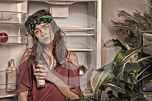 Woman with tired look standing next to open fridge. Girl holding glass of milk. Lady with curly hair and green headband