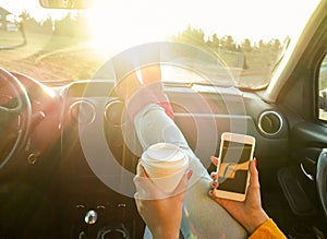 Woman toasting coffee take away go cup and using smart phone inside car with feet in warm socks on dashboard - Travel and trend