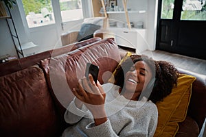 Woman with toothy smile lying on couch using smartphone at home listening to music on bluetooth wireless headphones
