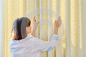 Woman touches curtains, lays out the folds on light velvet curtain