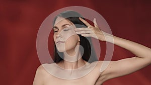 A woman touches the skin under her eyes, demonstrating the absence of wrinkles. Portrait of a woman on a red background