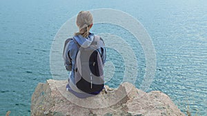 A woman tourist with a backpack on her shoulders sits on the top of a mountain and looks at the ocean