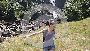 Woman tourist with backpack jogs on a path to rushing waterfall and raises hands up. Female backpacker runs on a trail