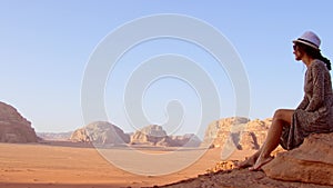 Woman tourist in dress sit on cliff barefoot at viewpoint on sunrise in Wadi Rum desert - valley Wadi Saabit. Jordan explore