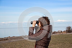 Woman tourist looking through binoculars at distant sea, enjoying landscape. Autumnal time.Lonely woman in brown coat