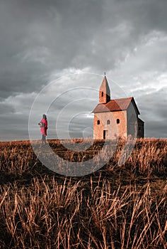 Žena turista v červeném kabátě stojící před starořímskokatolickým kostelem sv. archanděla michaela s temně zataženou oblohou v