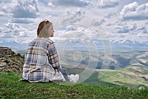 A woman tourist sits on the grass and looks at a beautiful landscape with a mountain range and a dramatic sky with