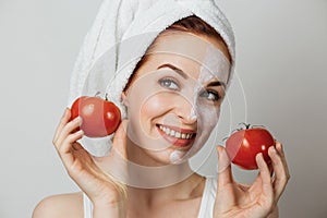 Woman in towel with cosmetic moisturizing mask on half of her face holding two fresh tomato.