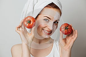 Woman in towel with cosmetic moisturizing mask on half of her face holding two fresh tomato.