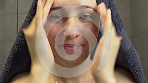 Woman with towel on her head apply skincare cream on her face and looking in mirror after shower. Female put moisturizing lifting