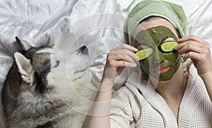 A woman with a towel on her head lies on a bed with a dog. on her face a green clay cosmetic mask with cucumbers. funny home spa