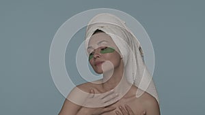 A woman with a towel on her head removes her hands from her face, opening it. A woman with patches under her eyes