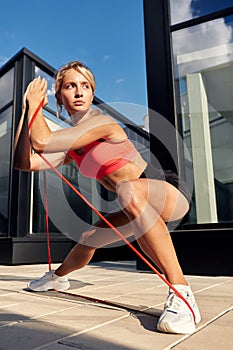Woman training outdoors with resistance band, doing squats, lunges, hip raises, positive girl in sportswear, in urban