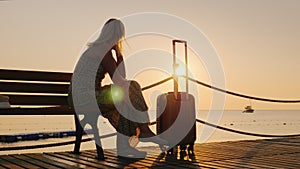 A woman with a travel bag sits on a wooden pier, looking forward to the dawn over the sea and a ship in the distance