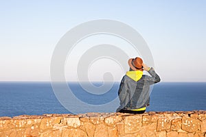 Woman in travel clothes and hat sitting and looking at blue ocean and sky