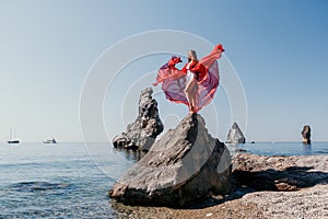 Woman travel sea. Young Happy woman in a long red dress posing on a beach near the sea on background of volcanic rocks