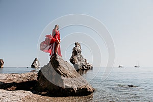 Woman travel sea. Young Happy woman in a long red dress posing on a beach near the sea on background of volcanic rocks