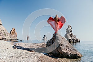 Woman travel sea. Young Happy woman in a long red dress posing on a beach near the sea on background of volcanic rocks