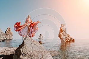Woman travel sea. Young Happy woman in a long red dress posing on a beach near the sea on background of volcanic rocks