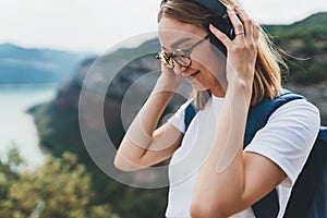 Woman traveler with backpack and hipster glasses listening to favorite music on wireless headphones while hiking on top of mount