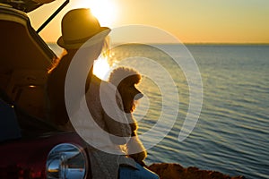 Woman traveler with dog sitting in car trunk near sea, watching sunset.