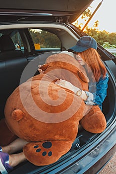 Woman traveler lying down on hatchback car and hugging a big bear