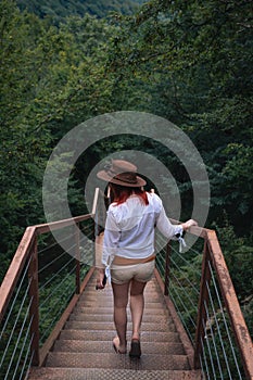 Woman traveler in Okatse Canyon in Georgia, standing on hanging metal pedestrian pathway trail above deep precipice. Leather hat.