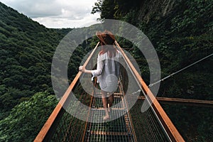 Woman traveler in Okatse Canyon in Georgia, standing on hanging metal pedestrian pathway trail above deep precipice. Leather hat.