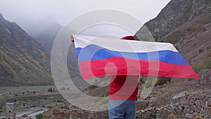 A woman traveler with a waving Russian flag on her shoulders in the mountains in autumn, enjoying the view of the misty
