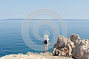 Woman traveler wearing straw summer hat and backpack, standing at edge of the rocky cliff looking at big blue sea and