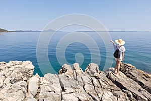 Woman traveler wearing straw summer hat and backpack, standing at edge of the rocky cliff looking and pointing at big